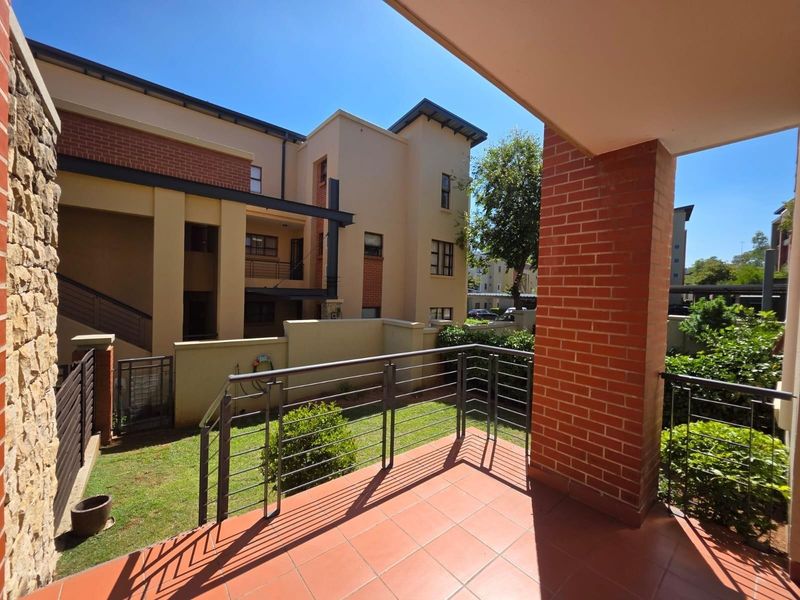 A covered patio with terracotta tiling and metal railings, overlooking a small private garden area within a modern apartment complex.