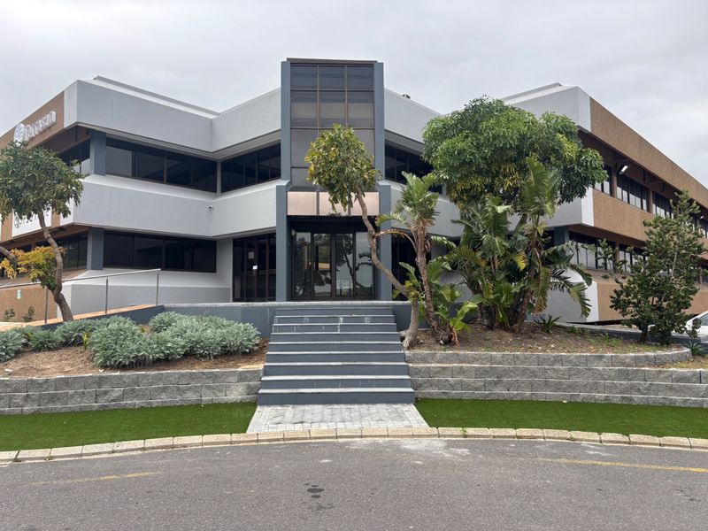 Front elevation of a modern multi-story office building featuring a central glass entrance, concrete retaining walls, and landscaped garden areas.