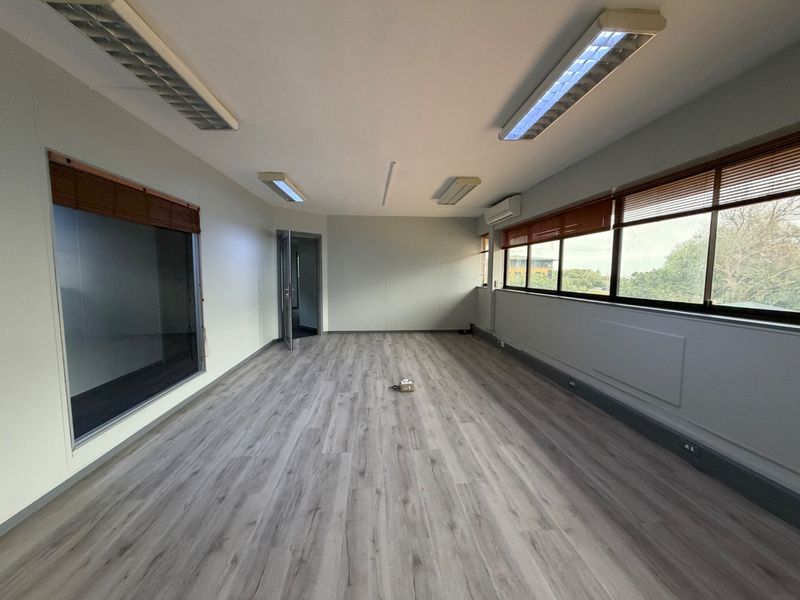 A bright, empty office space featuring modern laminate flooring, large windows with venetian blinds, and integrated fluorescent lighting.