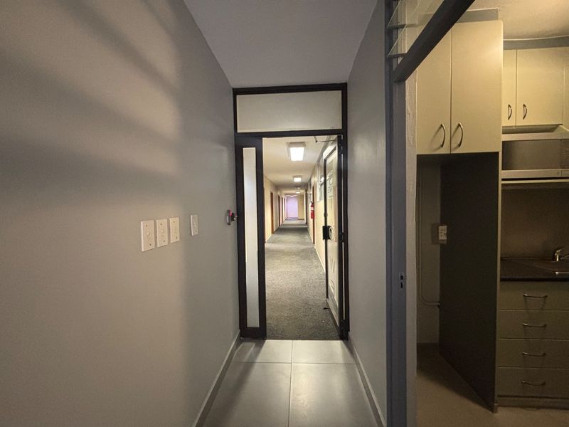 A functional office hallway featuring tiled flooring, leading to a long corridor with carpeted flooring and access to a small kitchenette area.