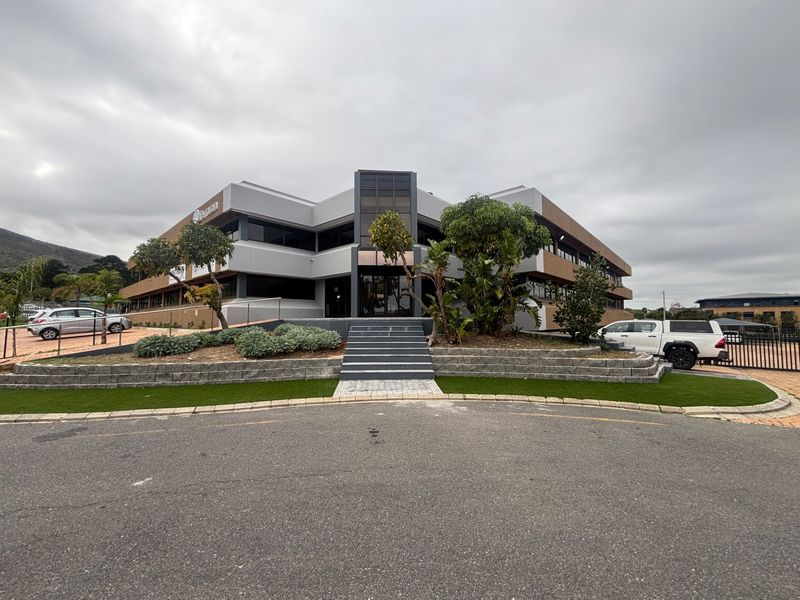 Front elevation of a modern multi-story office building featuring glass facade elements, paved entrance steps, and landscaped garden beds.