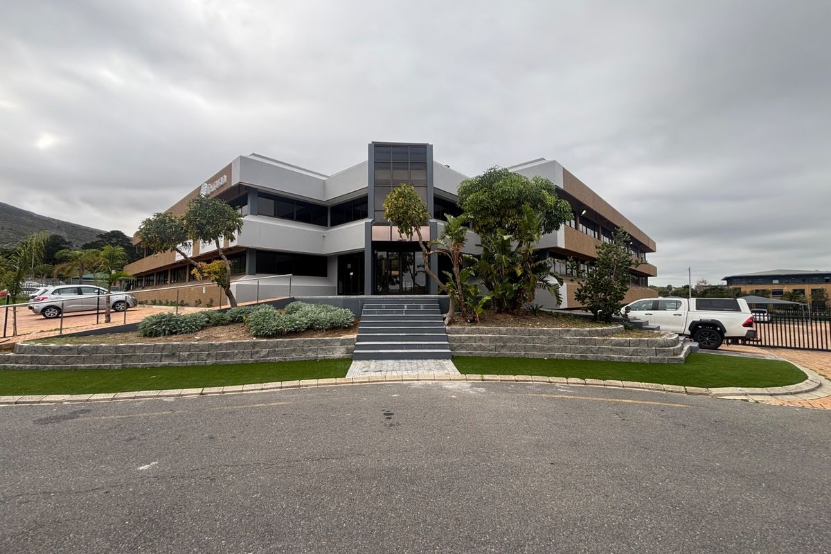 Front elevation of a modern multi-story office building featuring glass facade elements, paved entrance steps, and landscaped garden beds.