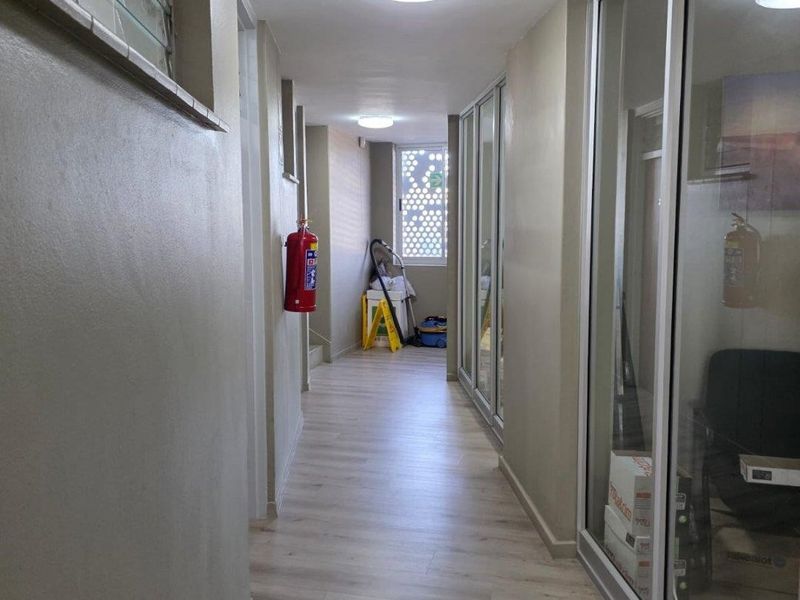 A well-lit commercial hallway featuring light wood-look flooring, glass-partitioned office walls, and mounted fire safety equipment.