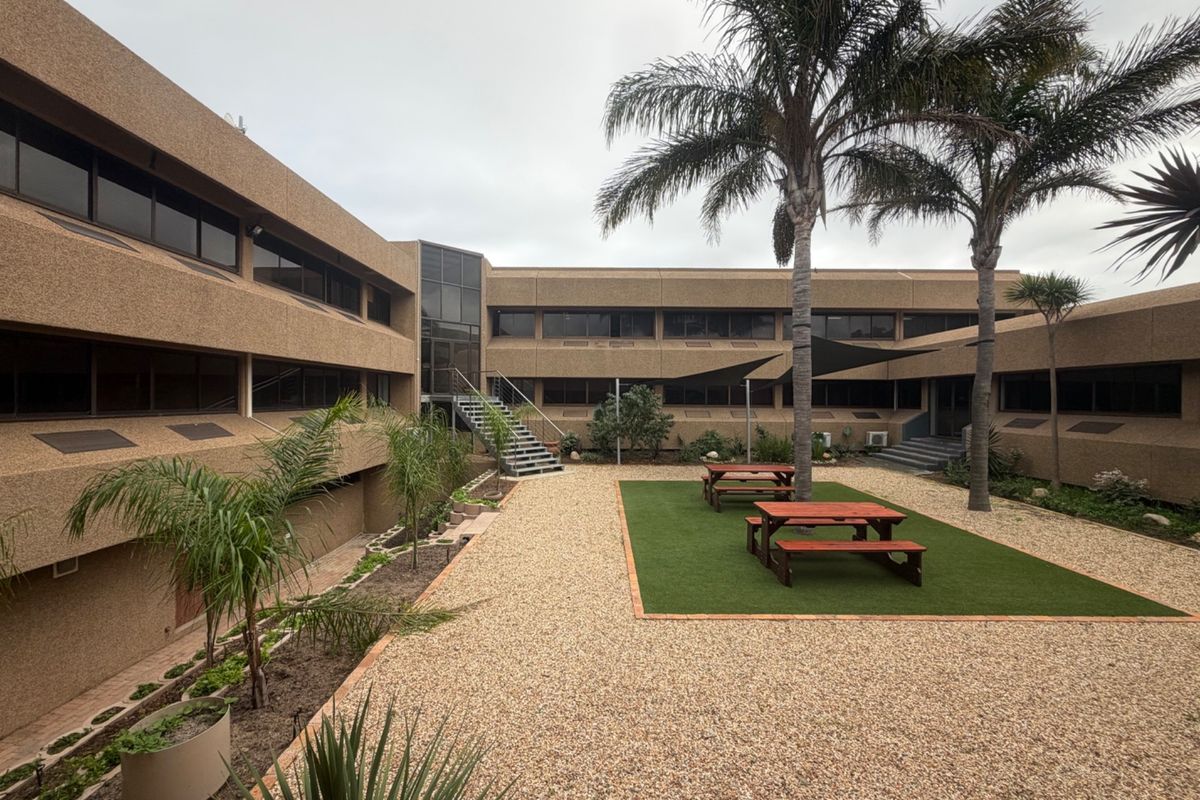 A well-maintained office courtyard featuring a pebble ground cover, artificial turf area, metal staircase access, and integrated shade sail for outdoor seating.