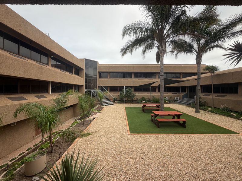 A well-maintained office courtyard featuring a pebble ground cover, artificial turf area, metal staircase access, and integrated shade sail for outdoor seating.