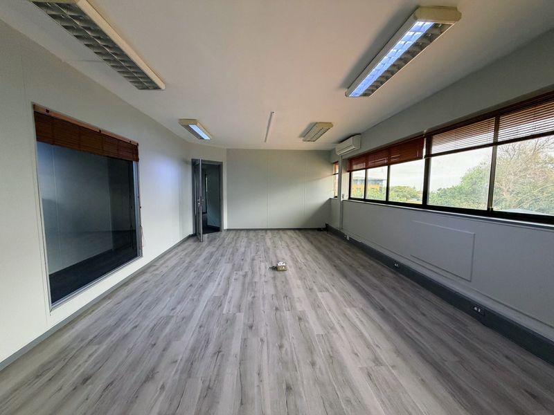 A bright, empty office space featuring modern laminate flooring, large windows with venetian blinds, integrated fluorescent lighting, and a wall-mounted air conditioning unit.