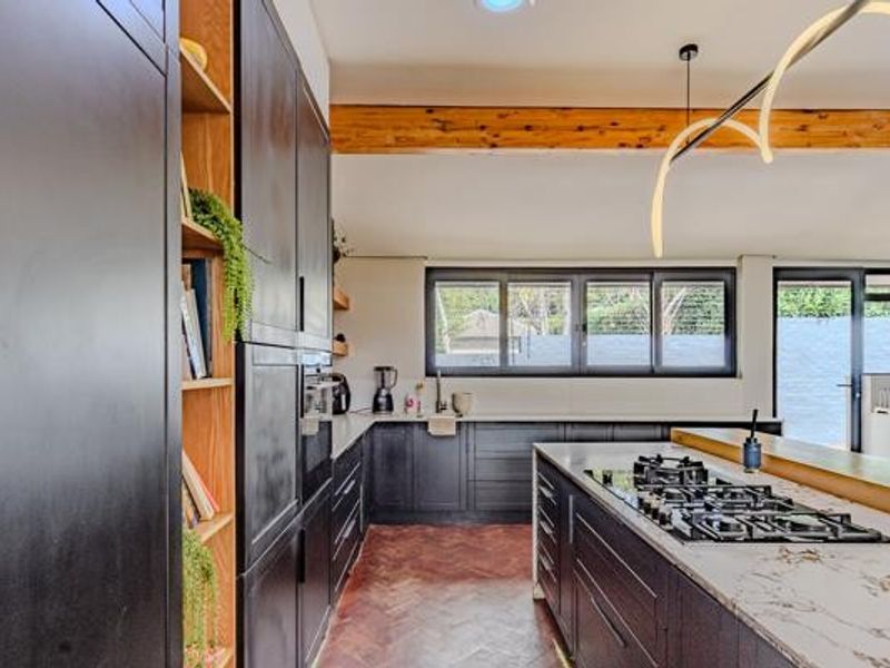 Modern kitchen featuring dark cabinetry, a central island with a gas cooktop, stone countertops, and elegant herringbone flooring under an exposed timber beam.
