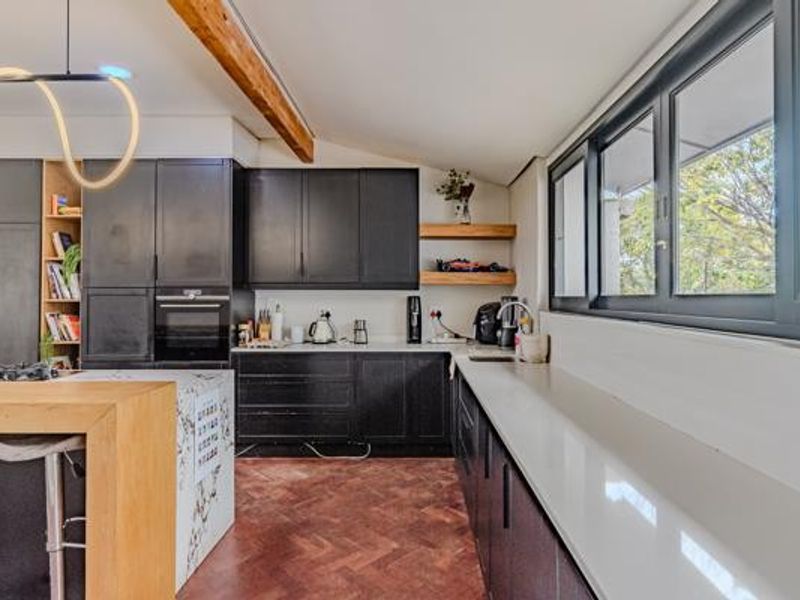 A modern kitchen featuring dark cabinetry, stone countertops, herringbone flooring, and an exposed timber beam, complemented by large windows and contemporary lighting.