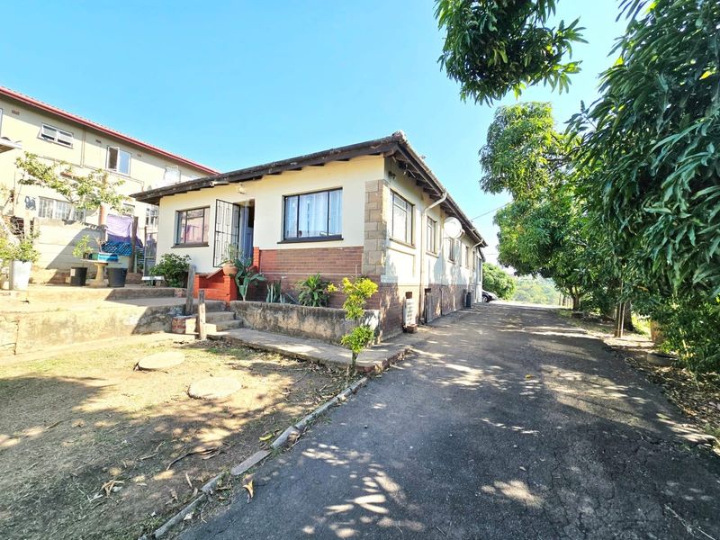 Exterior view of a single-story residential house featuring a brick and plaster facade, paved driveway, and surrounding garden area.