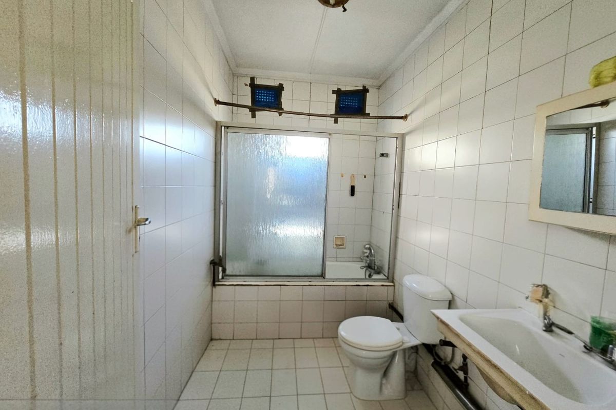 A dated bathroom featuring white wall and floor tiles, a built-in bathtub with a shower screen, a standard toilet, and a wall-mounted vanity basin.