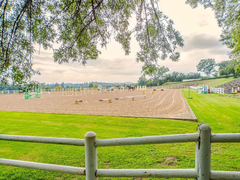 A well-maintained outdoor equestrian arena featuring a sand surface, jump obstacles, post and rail fencing, and tiered spectator seating.