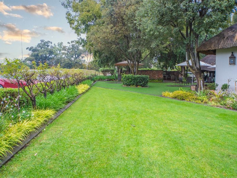 A well-maintained garden area featuring lush green lawns, mature trees, and a portion of a building with a traditional thatched roof.