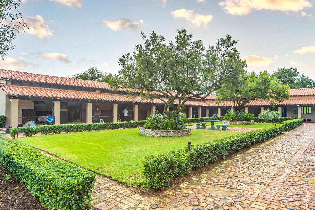 A well-maintained courtyard featuring a central lawn, mature trees, stone-edged garden beds, and a covered walkway with terracotta roof tiles.