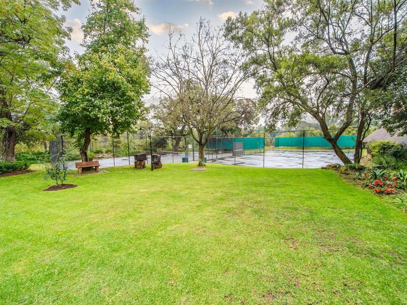 A well-maintained private tennis court set within a lush, mature garden environment, featuring perimeter fencing and established trees.