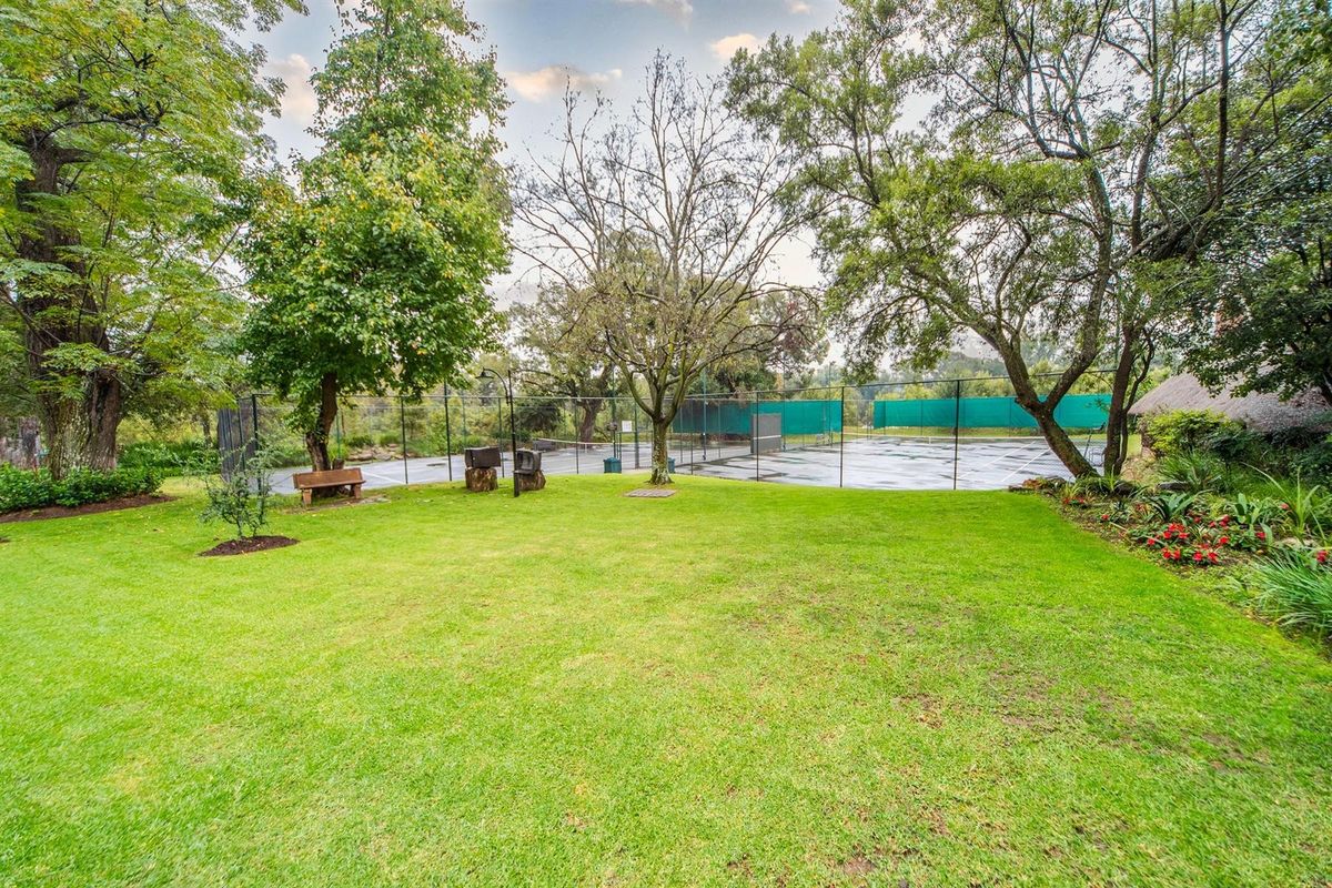 A well-maintained private tennis court set within a lush, mature garden environment, featuring perimeter fencing and established trees.