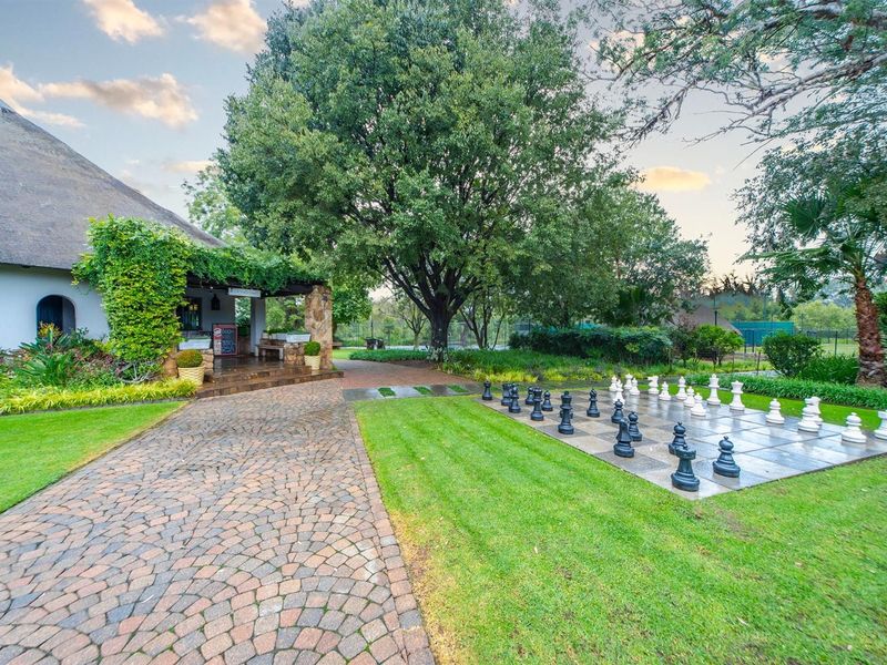 A well-maintained garden area featuring a stone-paved walkway, mature trees, and a permanent outdoor chess board set within a landscaped lawn.