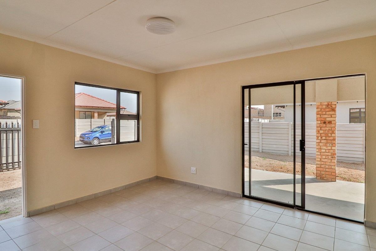 An empty living room featuring neutral tiled flooring, a sliding glass door leading to a patio, and large windows providing natural light.