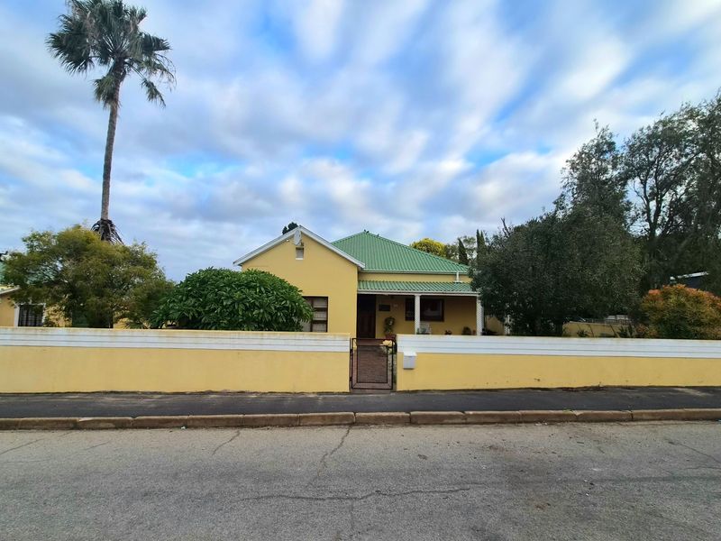 A charming single-story yellow house with a green metal roof, featuring a front boundary wall and gated entrance.