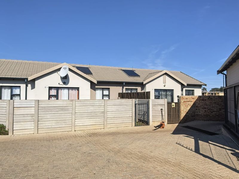 Exterior view of a townhouse complex featuring a paved driveway, concrete boundary wall, and tiled roof with solar panels.