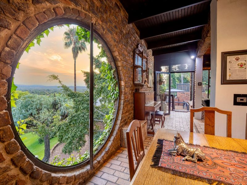 A unique dining area featuring a striking circular brick-framed window with expansive garden and sunset views, exposed brick walls, and timber ceiling beams.