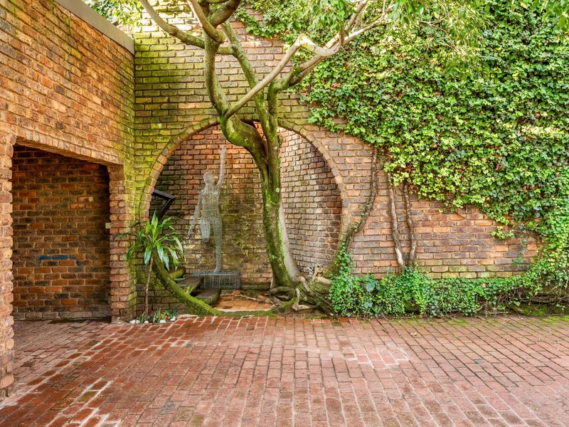A charming, rustic courtyard featuring exposed brick walls, a circular architectural archway, and mature tree integration with paved brick flooring.