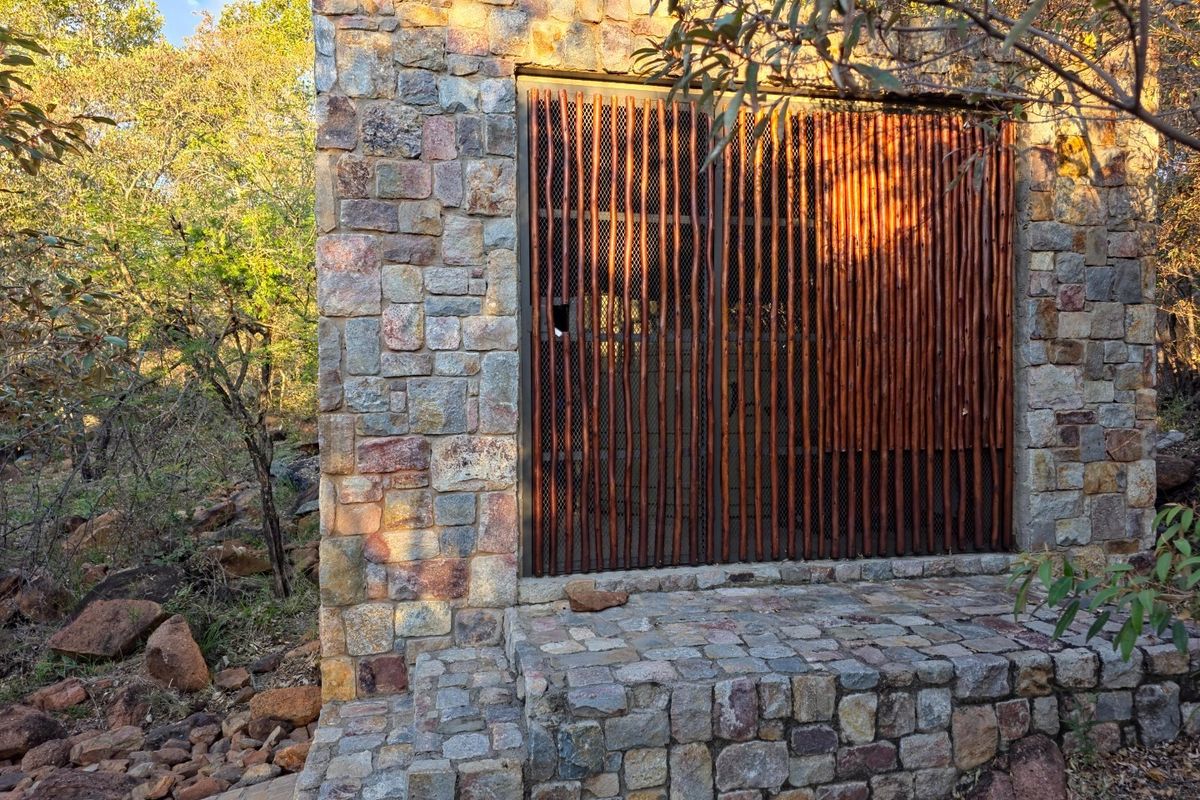 A rustic stone-clad exterior structure featuring a decorative timber slat screen and stone masonry steps, set within a natural bushveld environment.