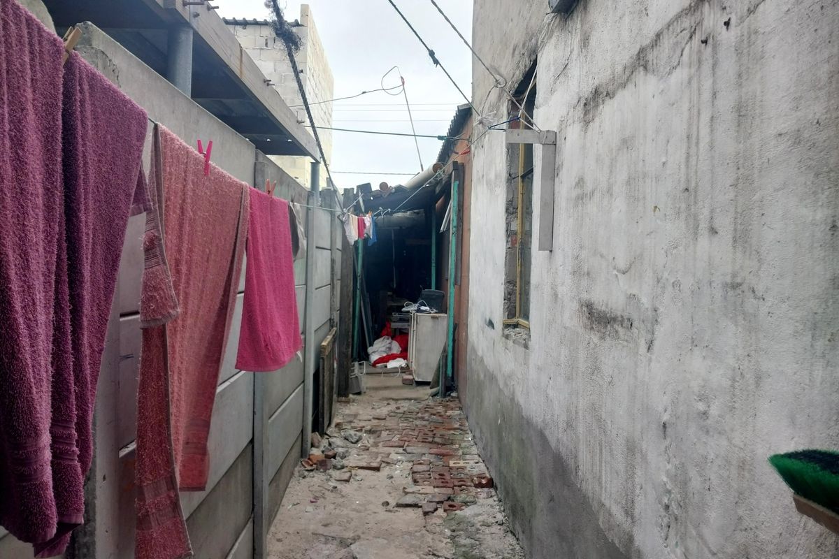 A narrow side alley or rear exterior passage featuring a precast concrete boundary wall and uneven brick paving in need of maintenance.