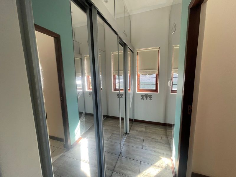 A bright bedroom featuring extensive built-in mirrored sliding wardrobes, tiled flooring, and windows fitted with roller blinds and security bars.