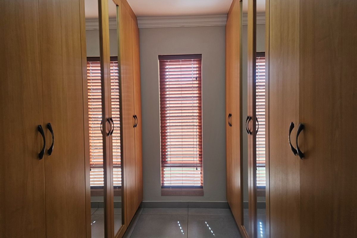 A well-maintained dressing area featuring floor-to-ceiling built-in wooden cupboards with integrated mirrors and a window with wooden venetian blinds.