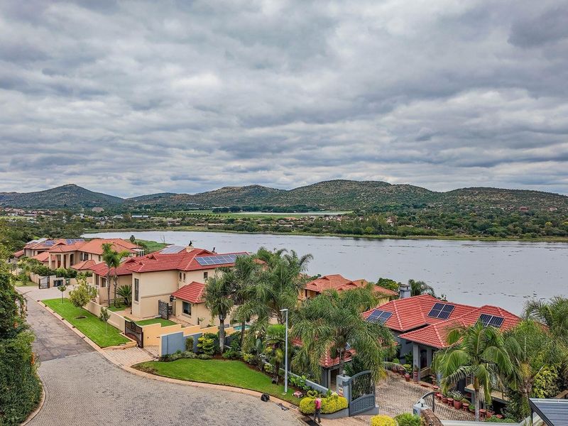 An elevated aerial view of a residential estate featuring homes with tiled roofs, solar panels, and paved driveways, overlooking a scenic lake and mountains.