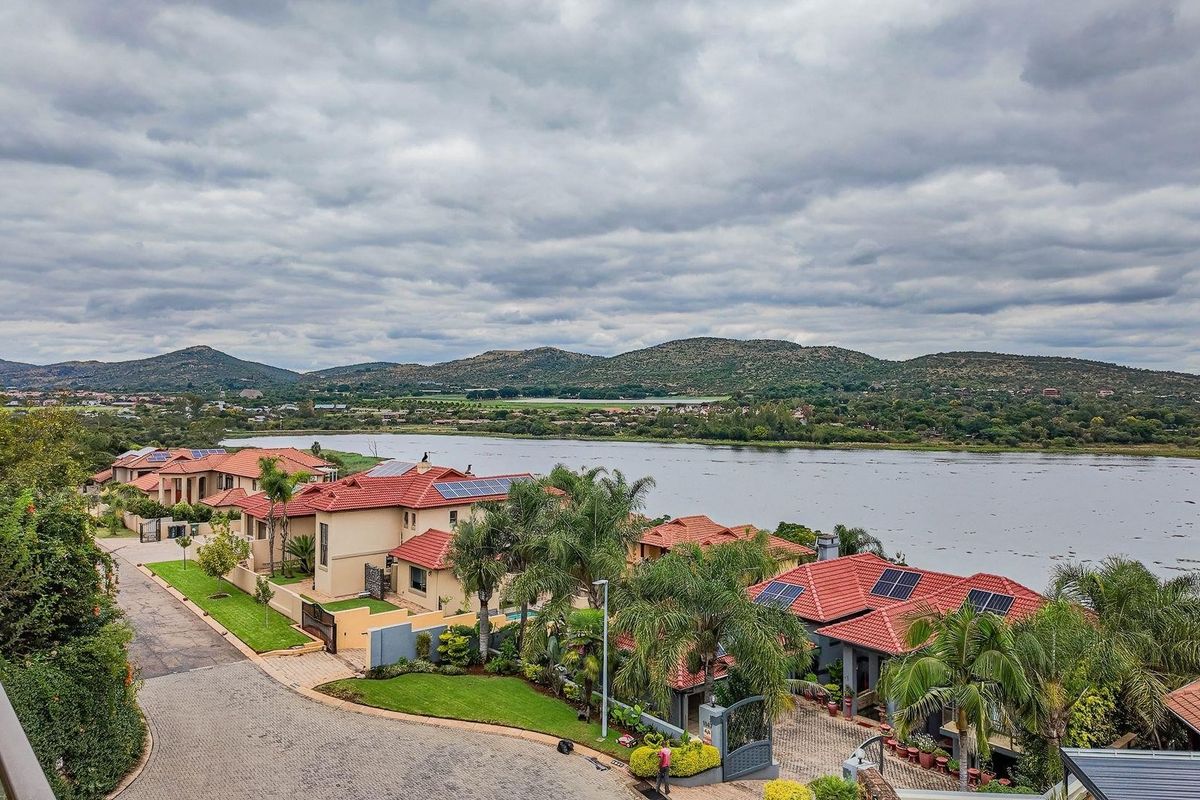 An elevated aerial view of a residential estate featuring homes with tiled roofs, solar panels, and paved driveways, overlooking a scenic lake and mountains.