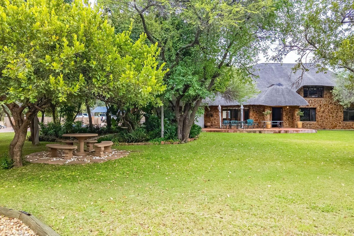 A spacious rear garden featuring a well-maintained lawn, mature trees, and a stone-clad farmhouse with a traditional thatched roof and covered patio area.
