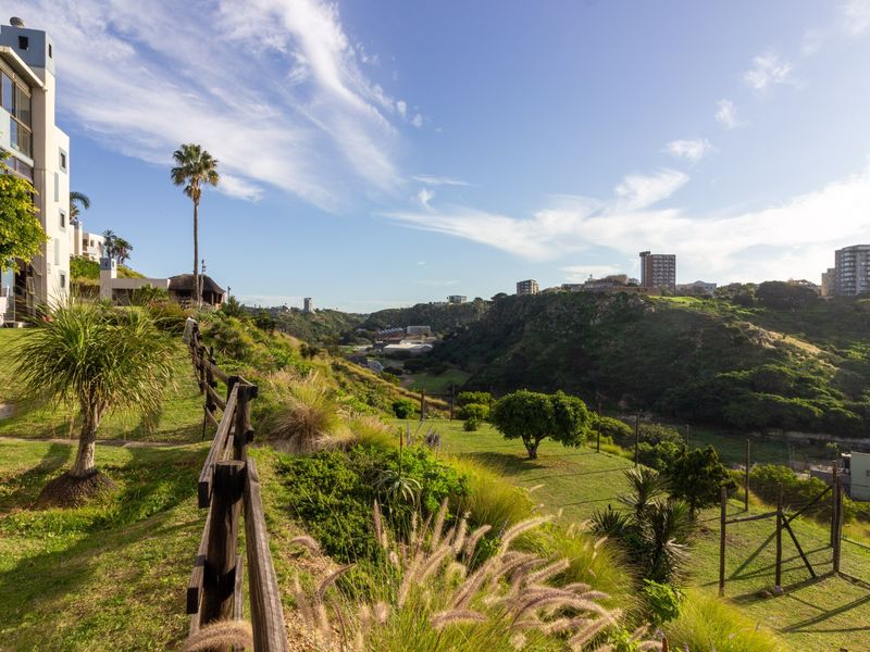 A scenic view overlooking a lush green valley and surrounding neighborhood, featuring a well-maintained garden area and wooden perimeter fencing.