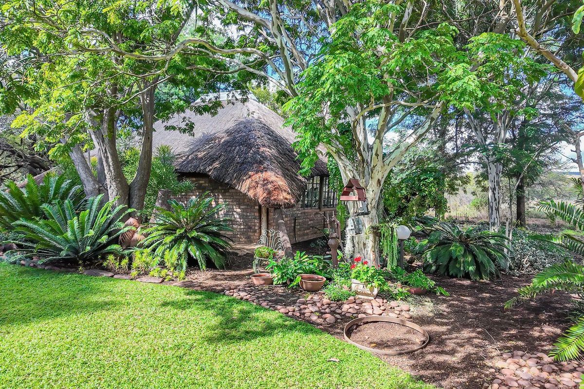 A view of a charming thatched-roof farm cottage nestled within a lush, mature garden setting with established trees and landscaped beds.