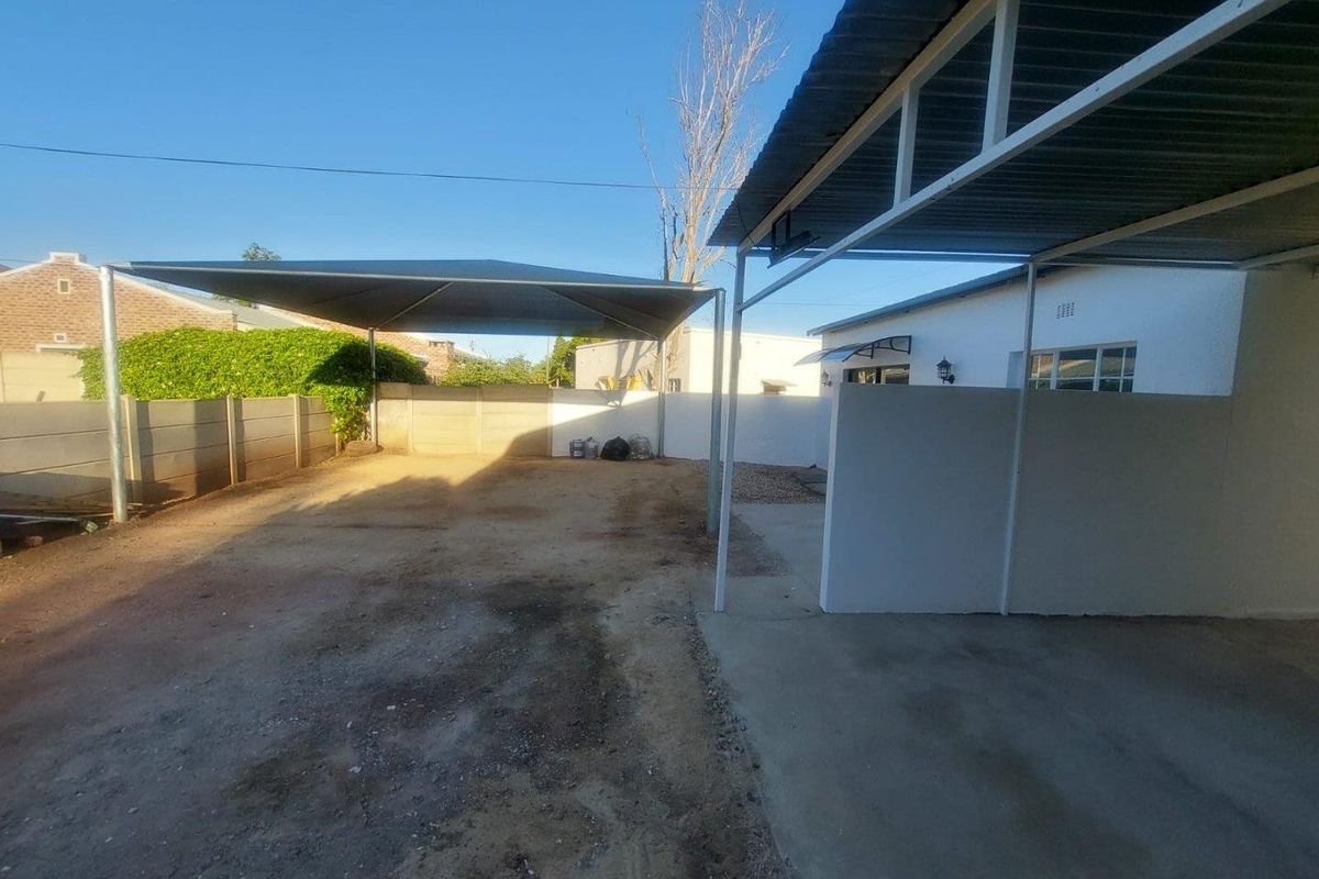 Outdoor area featuring a covered carport structure and a shade sail, providing sheltered parking space within a fenced property boundary.