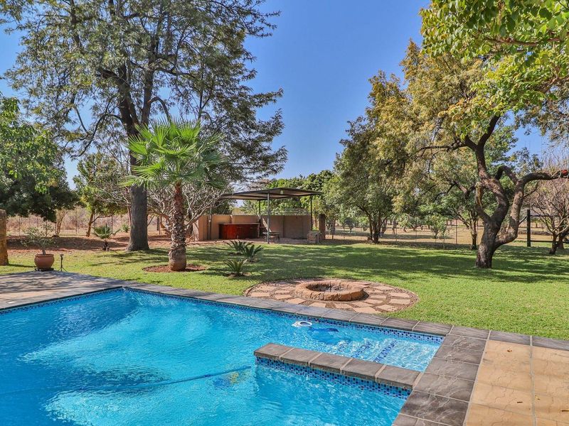 A well-maintained outdoor pool area featuring a tiled pool, stone-paved fire pit, and a covered entertainment structure set within a spacious, tree-lined garden.