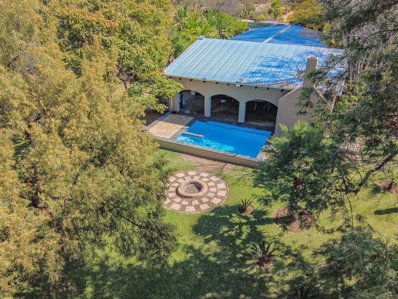 Aerial view of a farm property featuring a swimming pool, covered patio area, and a circular paved fire pit set within a landscaped garden.
