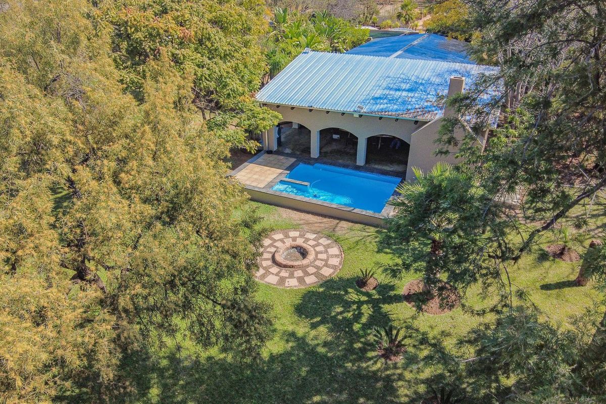 Aerial view of a farm property featuring a swimming pool, covered patio area, and a circular paved fire pit set within a landscaped garden.