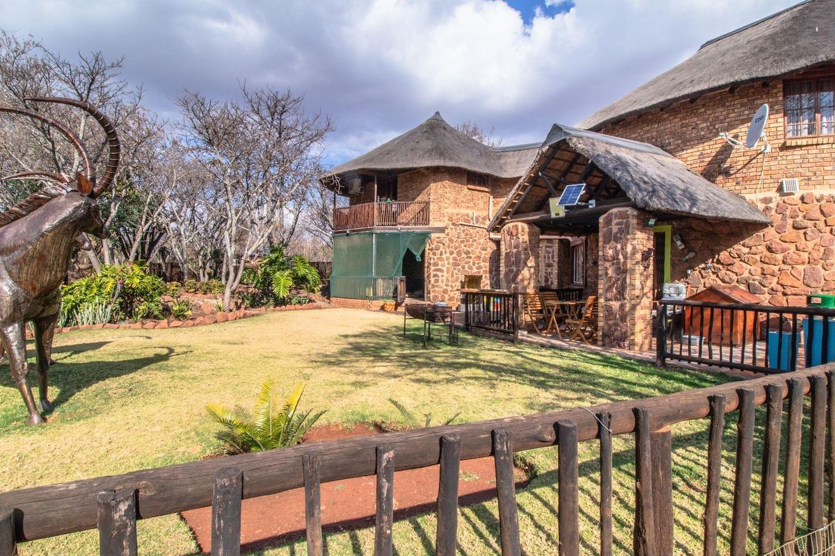 A spacious rear garden featuring a thatched-roof farmhouse, a covered patio area, and a large metal sculpture set within a landscaped lawn.