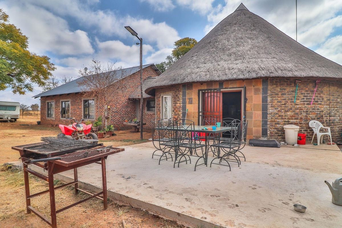 A rustic farm property featuring a traditional thatched-roof structure with an adjacent brick building and a spacious concrete patio area for outdoor living.