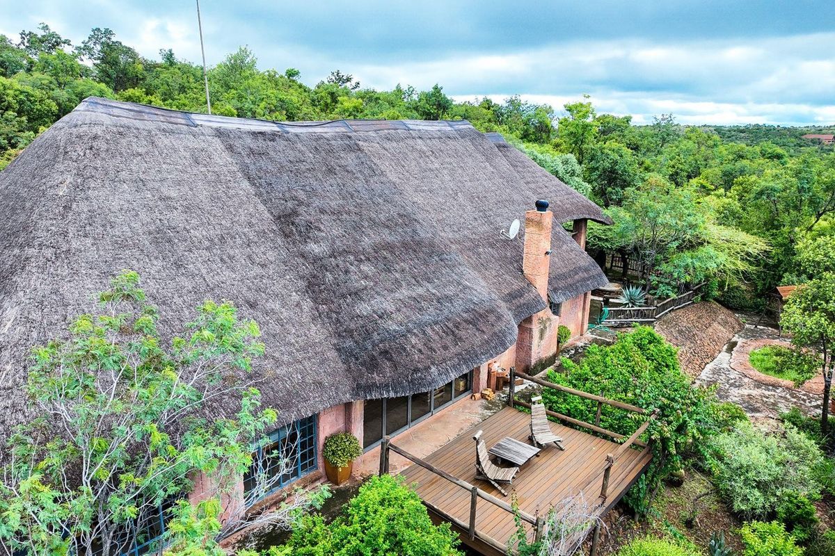 Aerial view of a farm property featuring a large thatched roof dwelling with an elevated wooden deck and surrounding natural landscape.