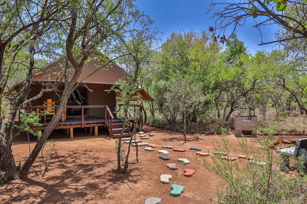 A rustic safari-style tented unit featuring a raised wooden deck, surrounded by natural bushveld, complete with an outdoor brick braai area.