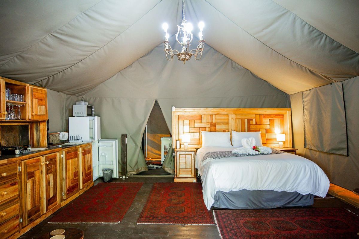 A luxury tented bedroom featuring a wooden headboard, built-in wooden kitchenette cabinetry, and a central chandelier within a canvas structure.