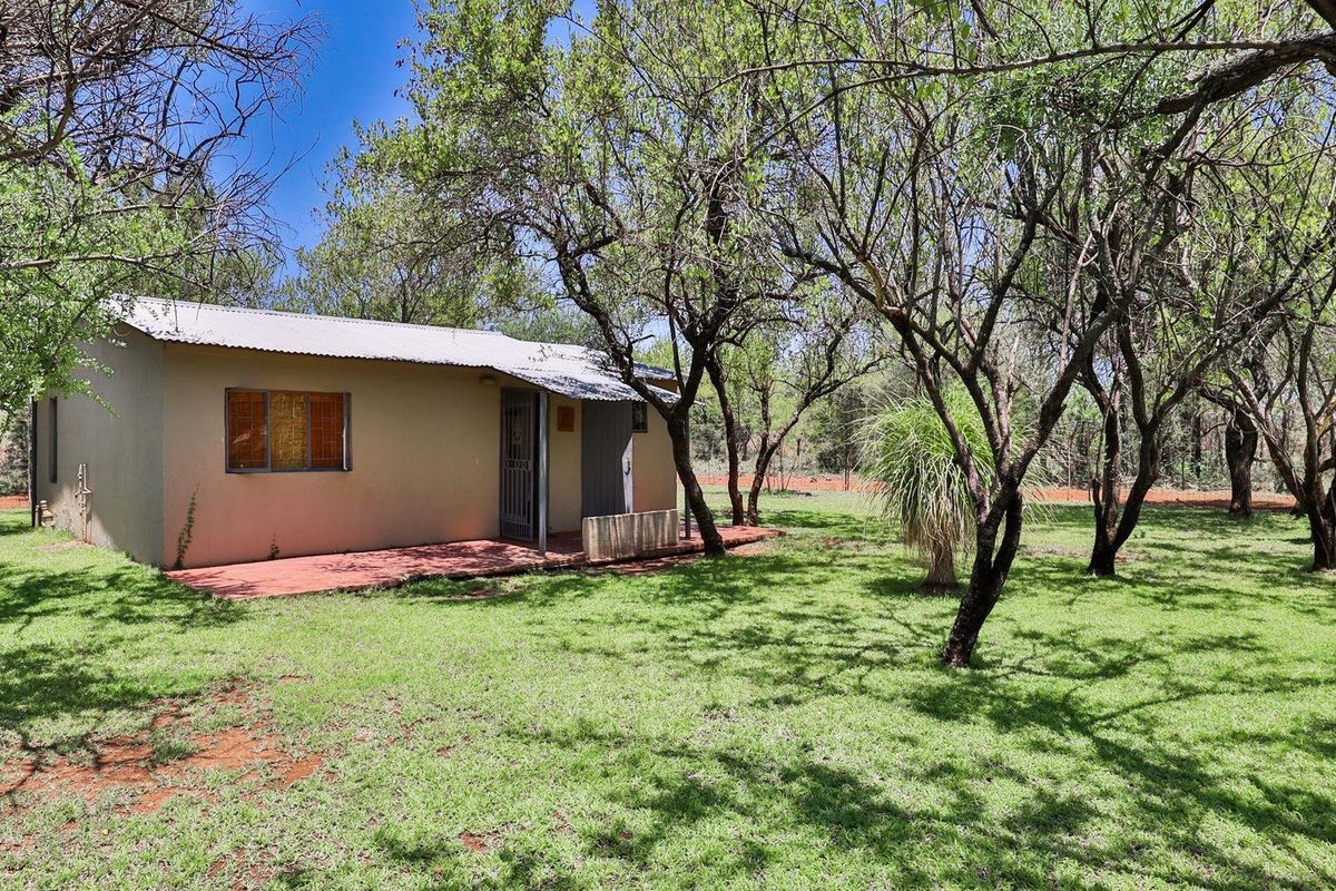 A modest farm cottage exterior featuring a simple facade, corrugated metal roof, and a small paved patio area set within a mature garden.