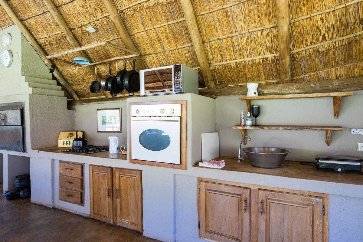 Rustic kitchen area featuring a built-in oven, gas hob, and basin, set under a traditional thatched roof with exposed timber beams.