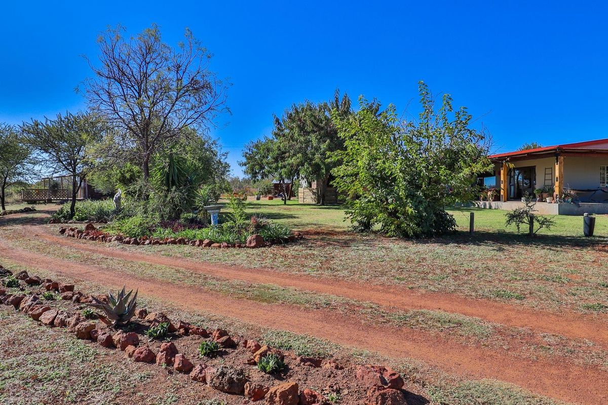 A wide view of the farm property featuring a dirt driveway, landscaped garden beds, and a covered patio area attached to the main dwelling.