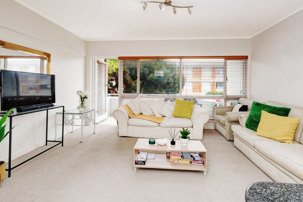 A bright living room featuring neutral walls, carpeted flooring, and large windows with blinds, providing access to an outdoor balcony area.