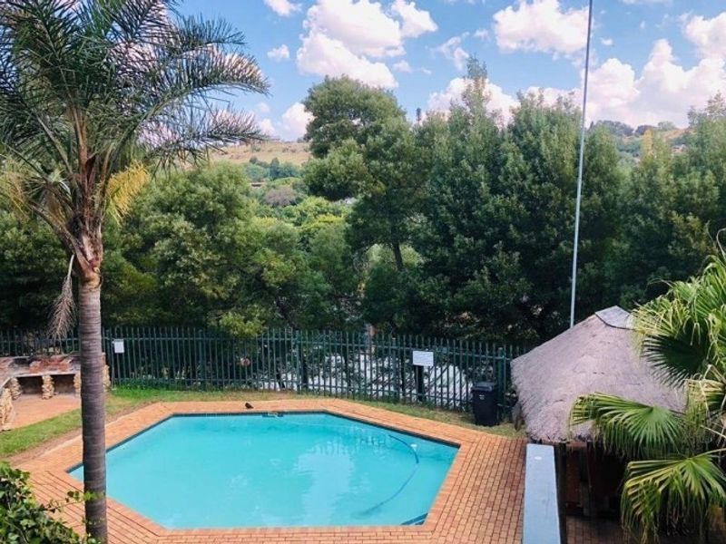 Outdoor pool area featuring a hexagonal swimming pool, brick paving, a thatched lapa, and a built-in stone braai area overlooking lush greenery.