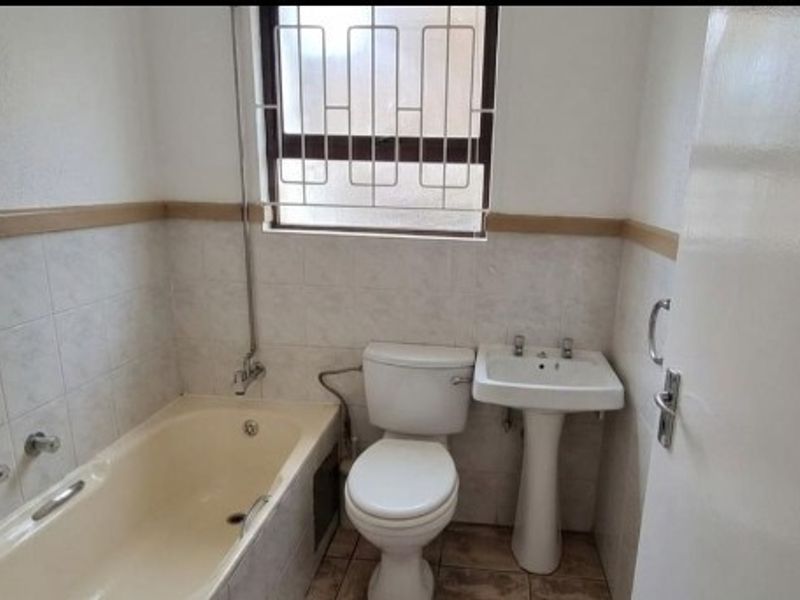 A compact bathroom featuring a standard bathtub, pedestal basin, and toilet, finished with neutral wall tiles and a security-barred window.