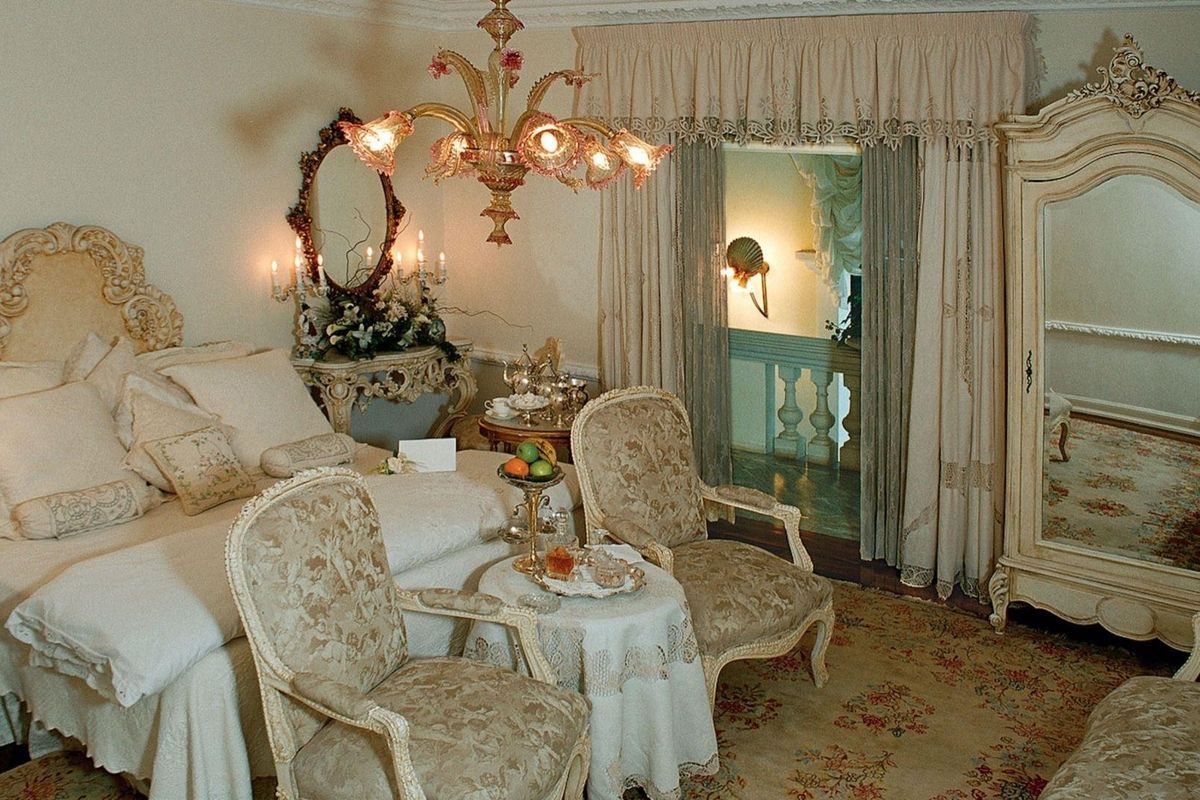 A classic bedroom featuring ornate ceiling molding, decorative wall sconces, and an open doorway leading to a balcony with a balustrade.